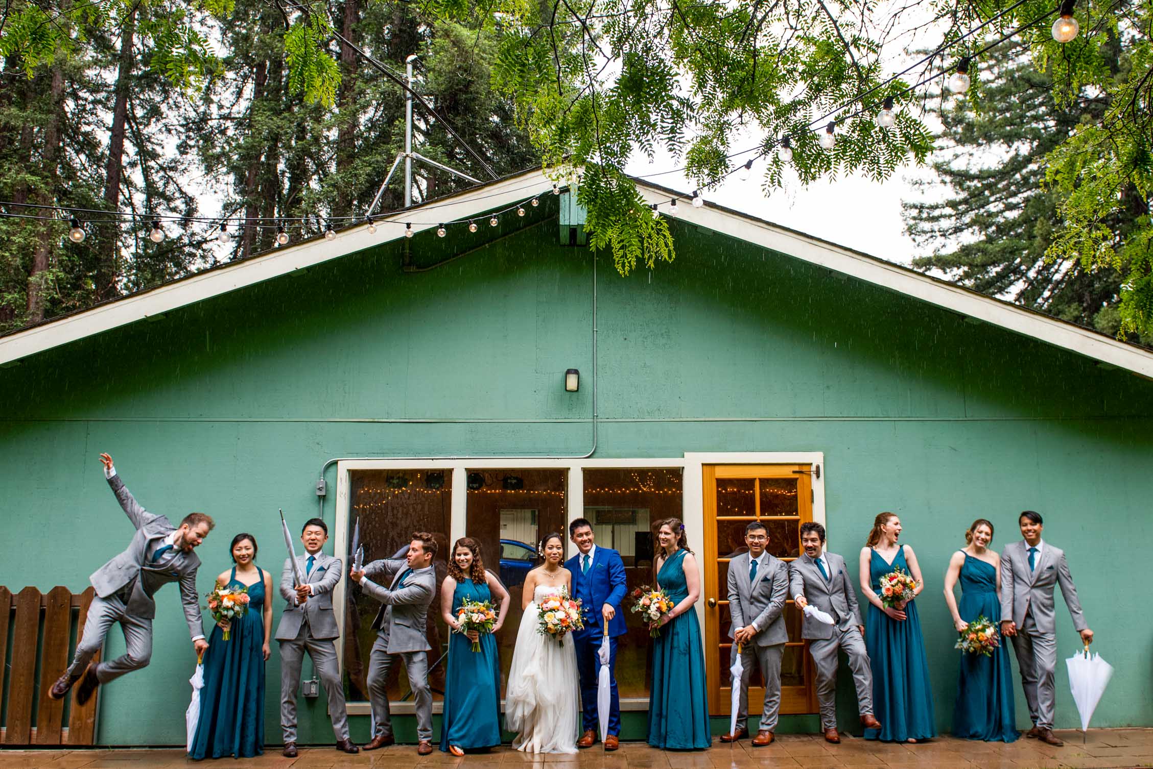 Large wedding party lined up under the awning of teal house with umbrellas