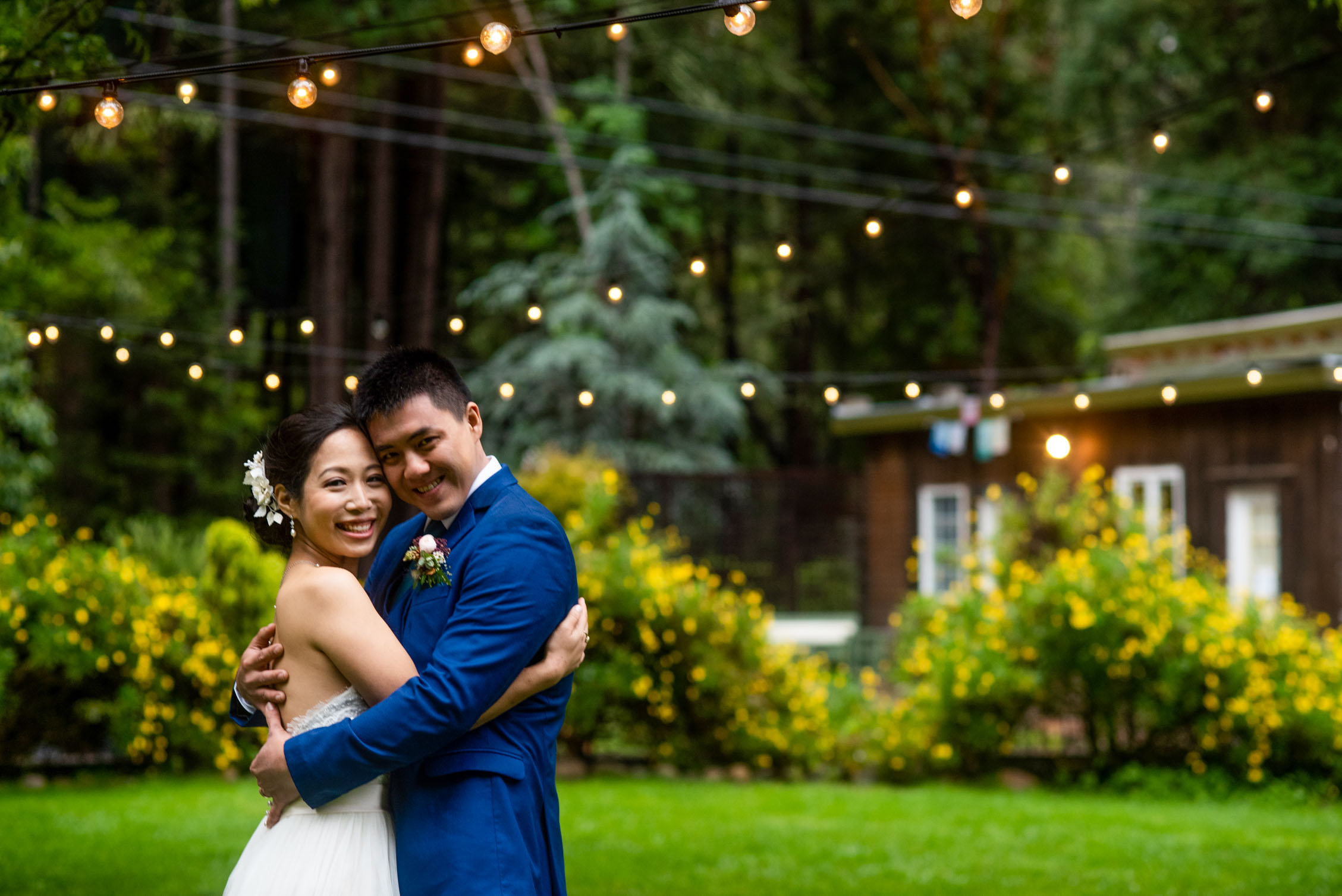 Bride and groom embrace on green lawn with string lights