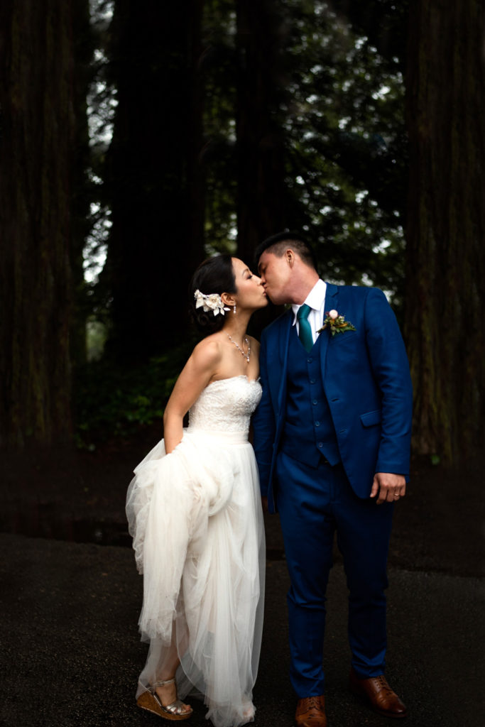 The bride holds her long white dress as she kisses her groom in the redwood forest